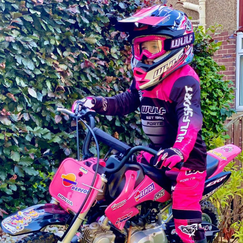 Girl sitting on a pink dirt bike with a pink and blue wulfsport suit and helmet